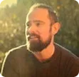 Bearded man with dark hair wearing a black shirt, smiling outdoors with sunlight in the background.